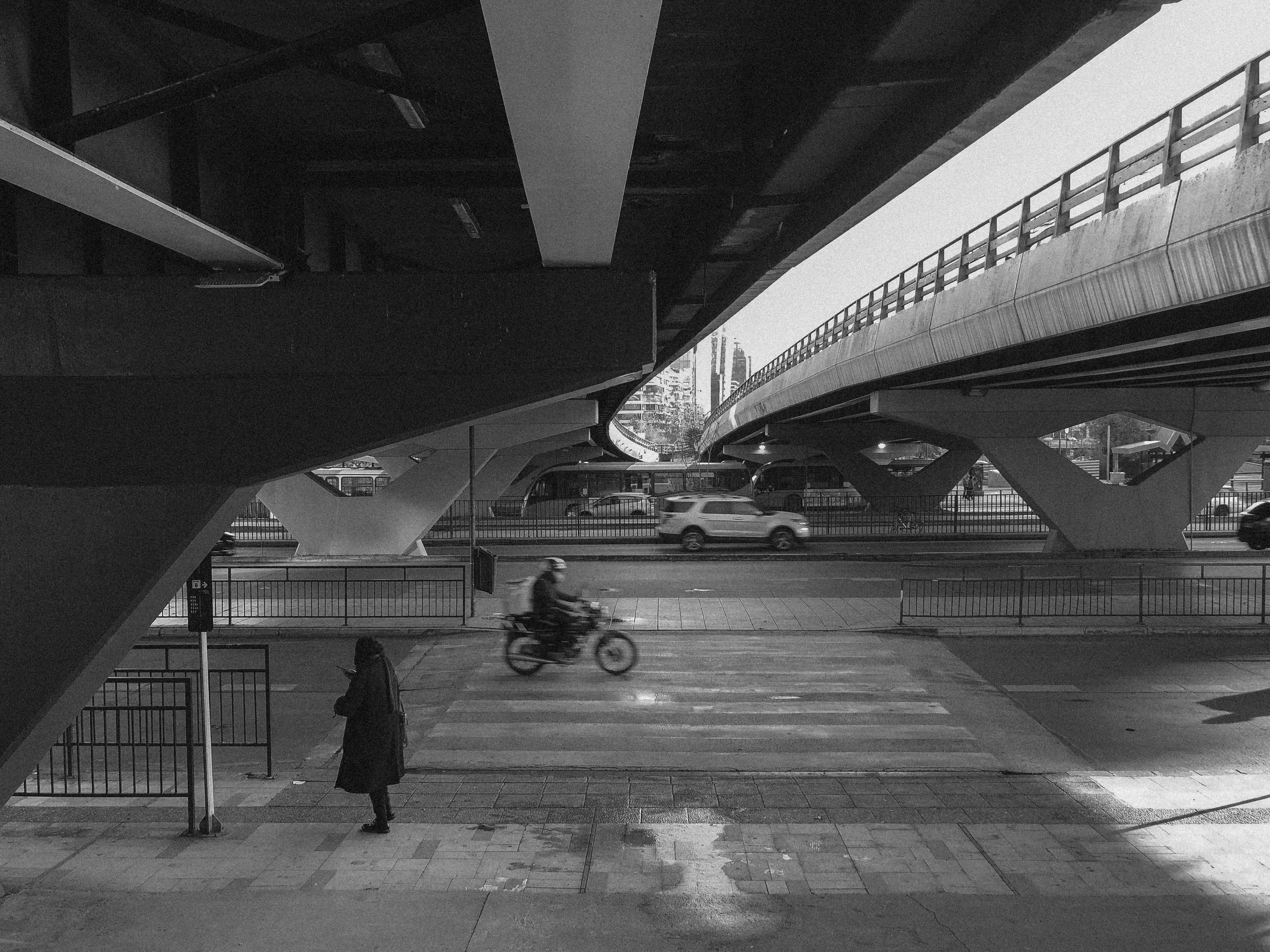 Black and white picture of an unknown woman standing alone in the bottom-left third of the image, surrounded by massive highways. Cars and motorcycles passing by. Noisy, low-contrast image.