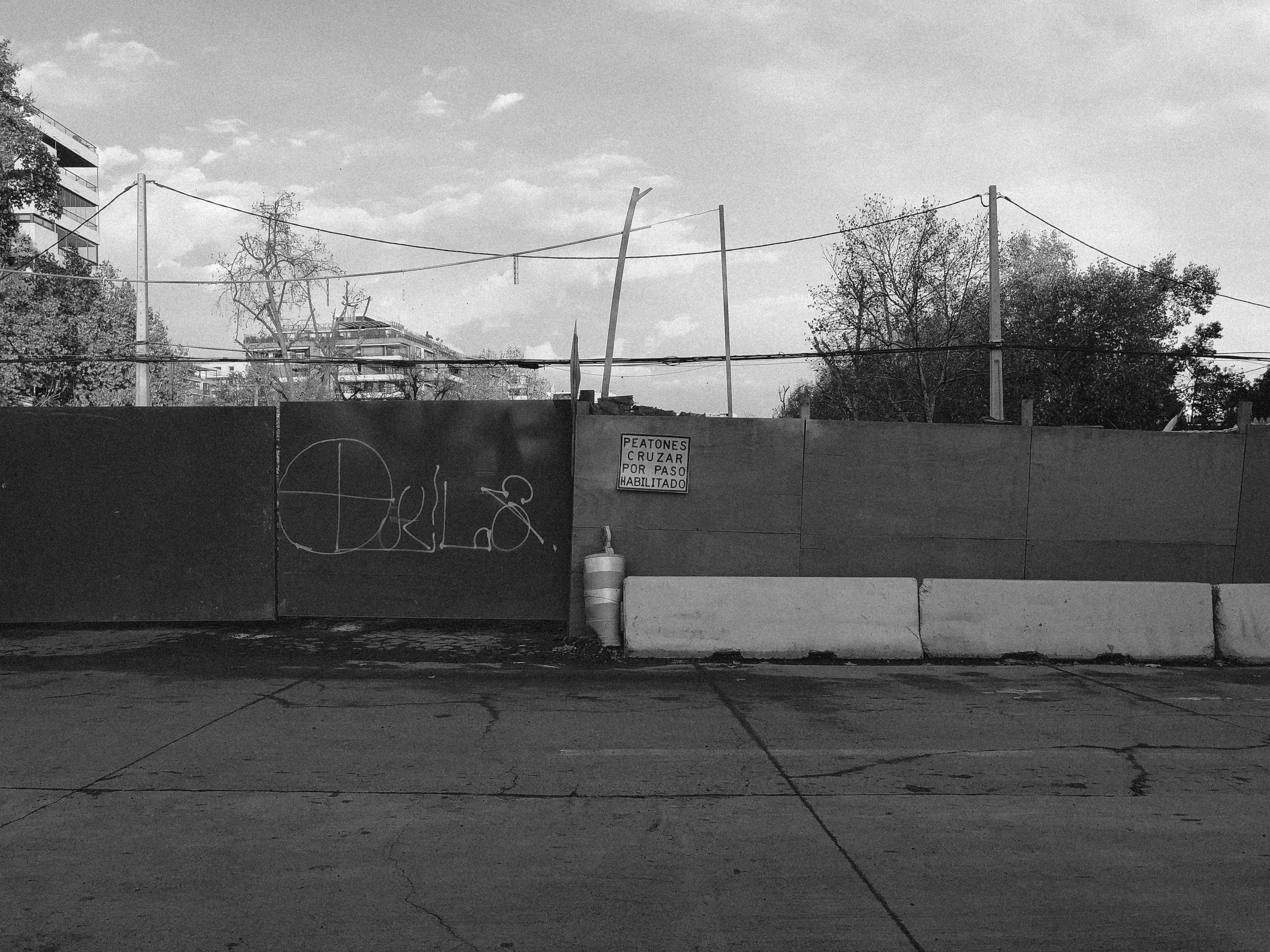 Black and white picture of an empty car passage, with the front of the pedestrian road blocked by giant panels and concrete structures. Hanged on the panels, a sign reads "Peatones cruzar por paso habilitado". The framing of the image conveys that the sign is blocking pedestrians from crossing by.